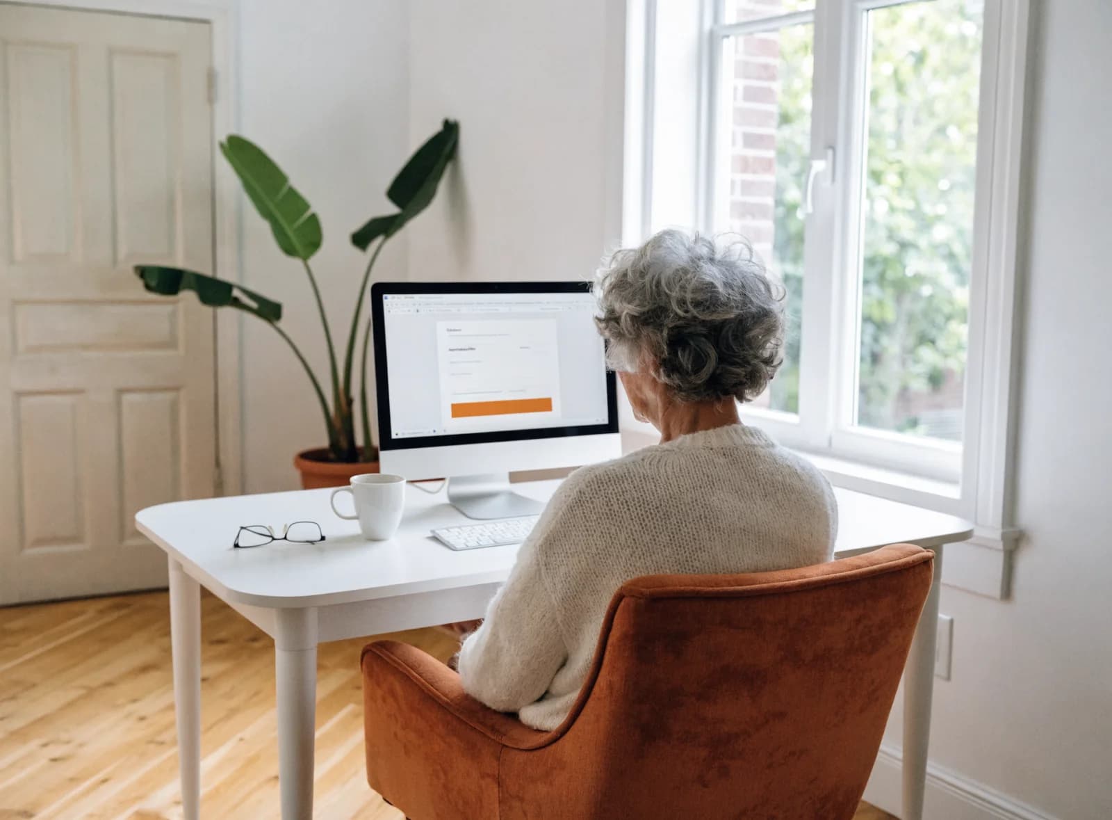 An older woman at a desktop computer in a bright living room, a clinic booking page on the screen